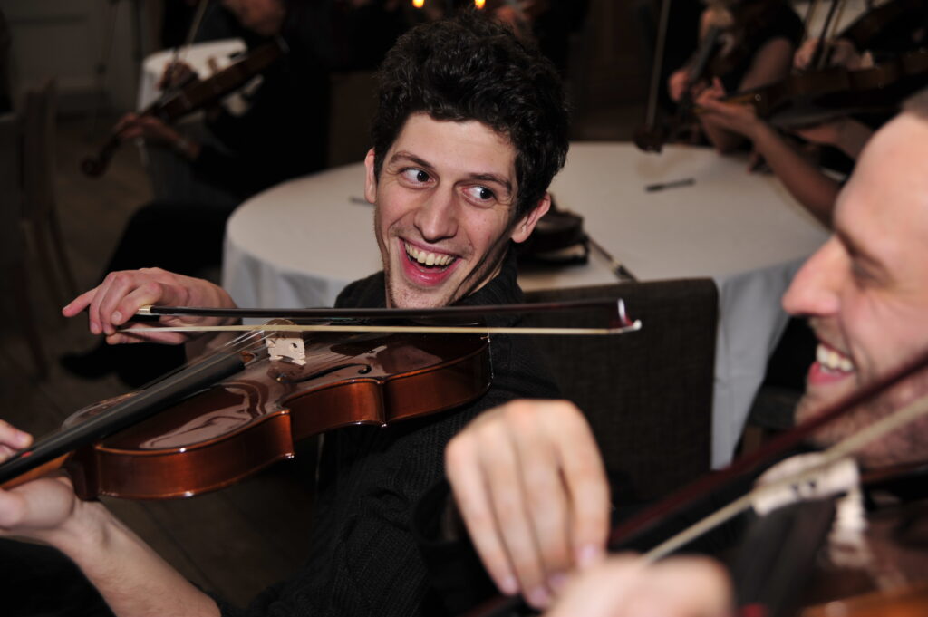 Two participants laughing and playing violins during the Crescendo team building event, creating music together in a joyful atmosphere.