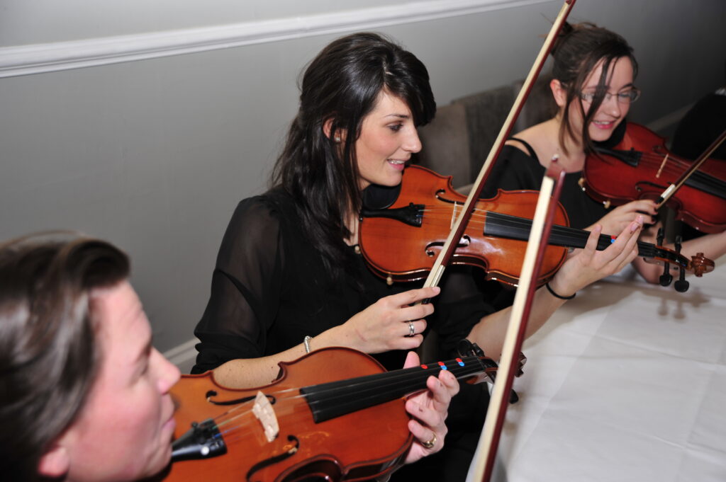 Participants smiling and playing violins together during the Crescendo team building event, highlighting teamwork and musical collaboration.