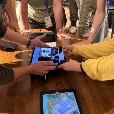 Participants in a 'Corporate CSI' team building event gather around a table, using iPads to examine digital evidence and clues as they work collaboratively to solve the mystery.