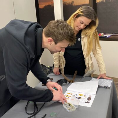 Two individuals are focused on assembling a prosthetic hand as part of The Prosthetic Hand Project. One person is intently working with components from a small plastic case, following the provided instructions laid out on the table. The second individual looks on, offering support and guidance during the assembly process. The setup on the table includes detailed assembly guides and various parts for the hand. This image captures teamwork, concentration, and the hands-on effort involved in building prosthetic hands during a team building program.