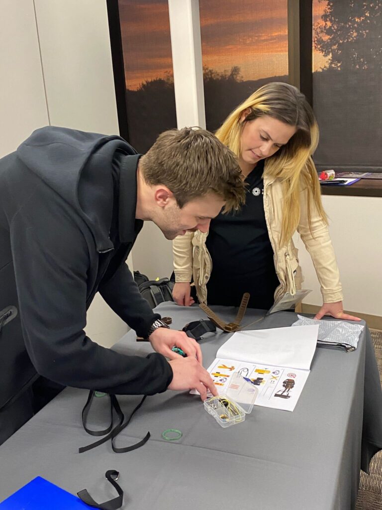 Two individuals are focused on assembling a prosthetic hand as part of The Prosthetic Hand Project. One person is intently working with components from a small plastic case, following the provided instructions laid out on the table. The second individual looks on, offering support and guidance during the assembly process. The setup on the table includes detailed assembly guides and various parts for the hand. This image captures teamwork, concentration, and the hands-on effort involved in building prosthetic hands during a team building program.