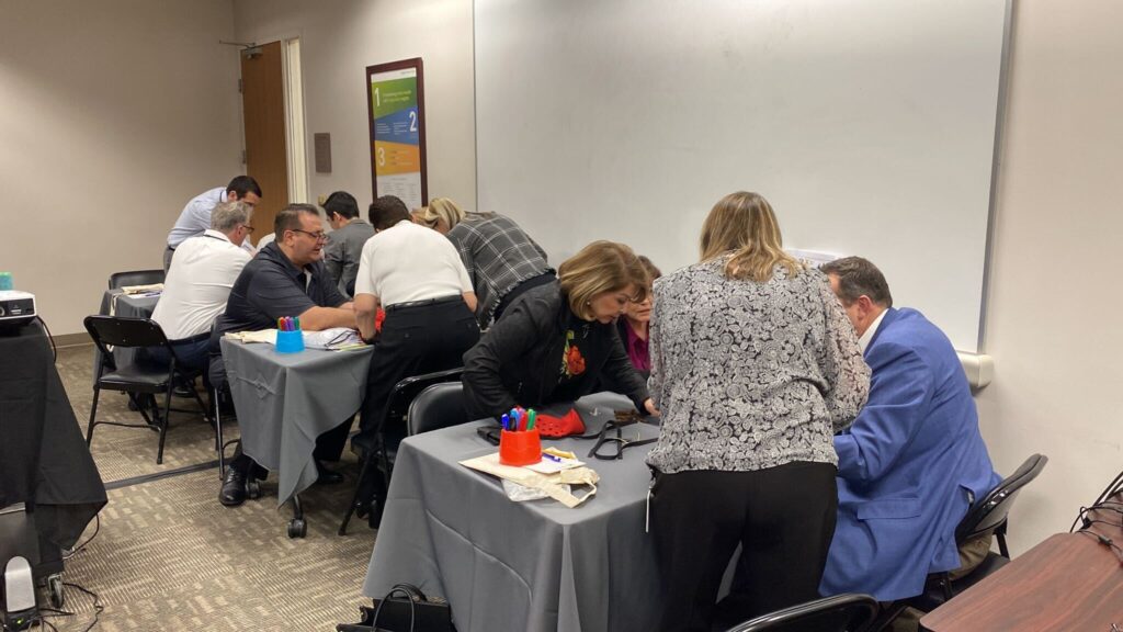 A group of individuals is seated at tables working on a team building activity in a conference room. Each table has participants intently focused on assembling prosthetic hands as part of a charitable event. Participants are wearing red empathy sleeves on their arms while carefully working with tools and parts.