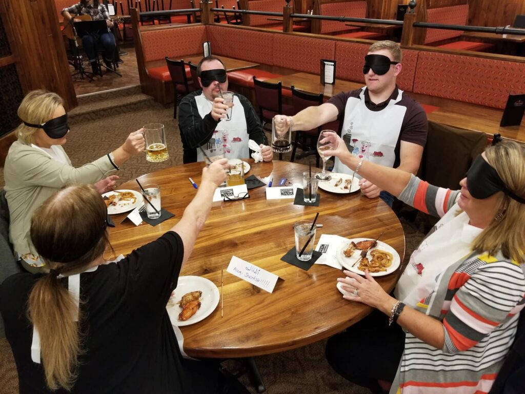 Group of participants blindfolded at a 'Dining In The Dark' team building event, toasting with drinks around a wooden table, enhancing trust and communication skills.