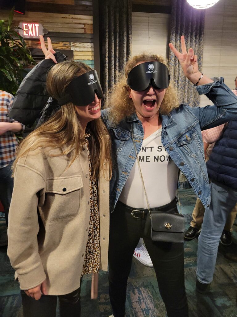 Two women wearing blindfolds, smiling and making playful gestures during a team building activity, creating a fun and engaging atmosphere.