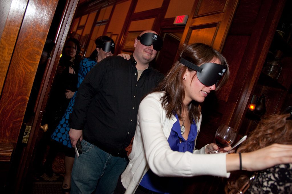 Participants navigating a Dining in the Dark event, wearing blindfolds and engaging with each other while exploring their senses and teamwork during a unique dining experience.