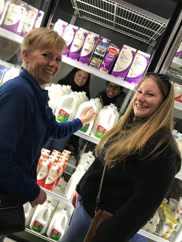 Four women are smiling as they stock or organize shelves in a refrigerated section of a grocery store, holding large containers of milk. Two women are in front, with two more standing inside the fridge behind them, all happily engaged in a Do Good Bus team building program.