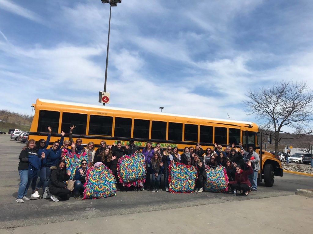 A large group of participants posing in front of a bright yellow school bus after a Do Good Bus event. The group, consisting of around 30 individuals, proudly holds colorful handmade blankets, a result of their charitable work. They stand together in a parking lot under a blue sky, smiling and waving to the camera. The Do Good Bus program encourages team building through mystery volunteer projects, helping communities in a fun and engaging way.