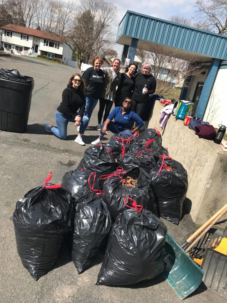 A group of six women poses proudly next to a collection of large black trash bags filled with yard waste and debris during a team building clean-up event. The women are casually dressed and smiling, indicating their sense of accomplishment.