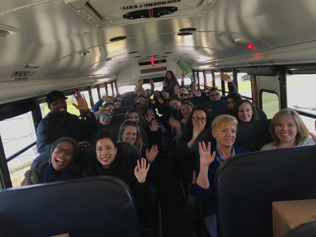 A group of smiling participants is seated inside a school bus, waving at the camera. The bus is well-lit with natural light, and the energy of the group is lively, as they seem ready for or returning from a team building event. The mixture of cheerful expressions and raised hands reflects a sense of fun, camaraderie, and excitement.