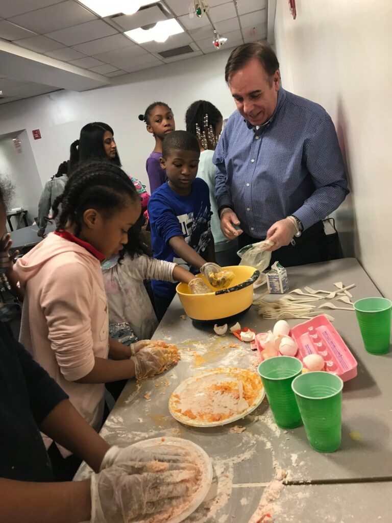 A group of children and an adult are engaged in a cooking activity. The children, wearing gloves, are mixing ingredients in a large yellow bowl while the adult, dressed in a blue shirt, assists them. The table is cluttered with ingredients, including cracked eggs, mixing bowls, and a plate with food remnants. The scene is lively, with other children in the background observing or waiting their turn to participate. The activity promotes teamwork and hands-on learning, part of a community team building event.