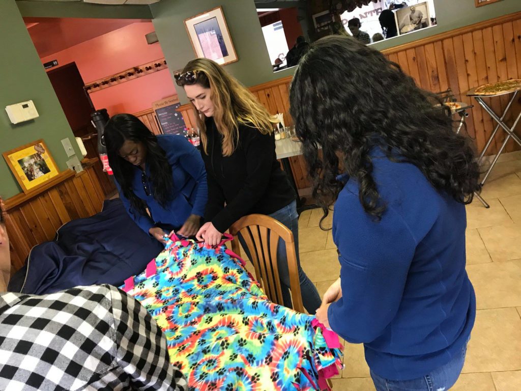 Three women are focused on crafting a colorful, tie-dye fleece blanket during a community team building project. The blanket features bright rainbow patterns and is being assembled on a wooden table. The women, casually dressed, are working together to tie the edges of the fabric, demonstrating teamwork and collaboration. Additional people and materials are partially visible in the background, contributing to the sense of a group project in progress.