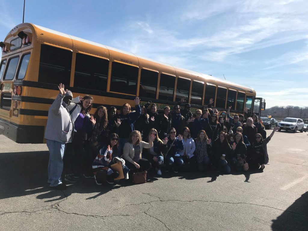 A large group of people poses in front of a yellow school bus on a sunny day, many of them waving at the camera. They are outdoors in a parking lot, with clear skies above, creating a sense of enthusiasm and camaraderie. The group appears to be preparing for or returning from a volunteer outing, part of a team building event. The mix of smiles and waves highlights the positive energy and collaborative spirit of the group.