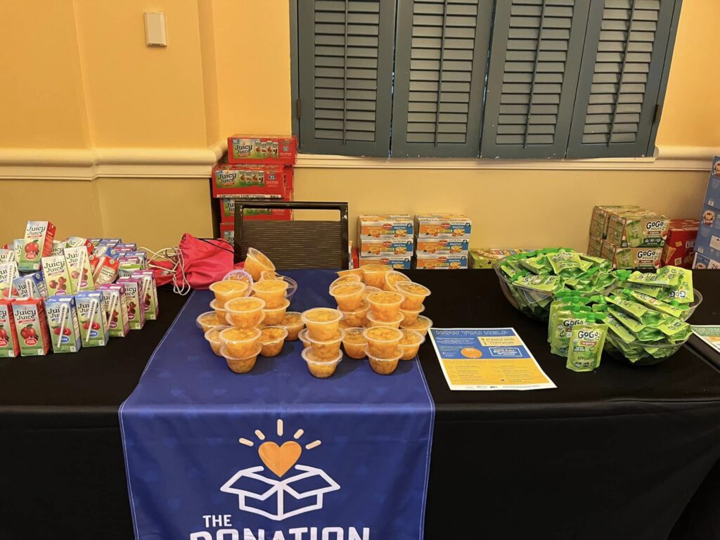 Table filled with fruit cups, juice boxes, and other snacks, set up for The Great Snack Pack activity at a corporate charitable team building event, with The Donation Station banner displayed in front.