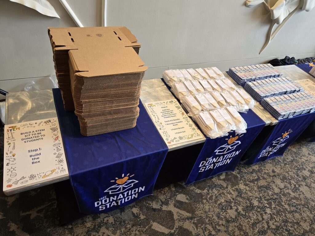 Table set up with flat cardboard boxes, STEM kits, and educational materials, ready for assembly during the Give the Gift of STEM corporate team building event, with The Donation Station banners displayed.