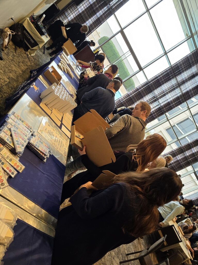 Participants assembling STEM activity kits in a line at a table covered with The Donation Station banner during the Give the Gift of STEM corporate team building event'.