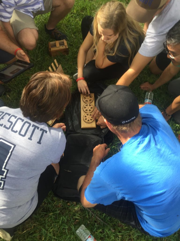 Team members collaborate outdoors on a gear puzzle during the 'Escape The Maze' team building activity, strategizing together to solve the challenge in an engaging hands-on exercise.