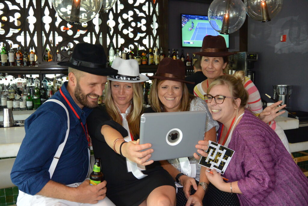 Group of five people in hats smiling and looking at a tablet during a team building activity at a bar, participating in the 'Escape the Mob' program. They are engaged in solving a scavenger hunt and murder mystery challenge, creating a fun and interactive atmosphere.