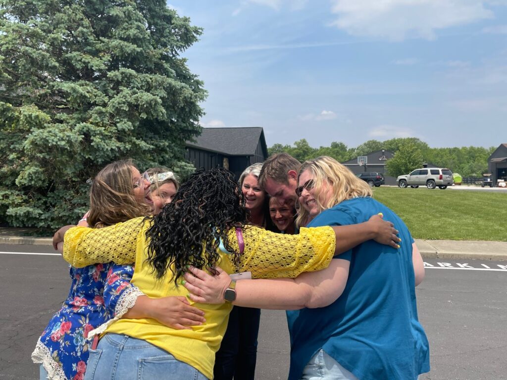 A group of six teammates sharing a heartfelt group hug outdoors during a team building event, standing on a paved road with greenery and trees in the background.