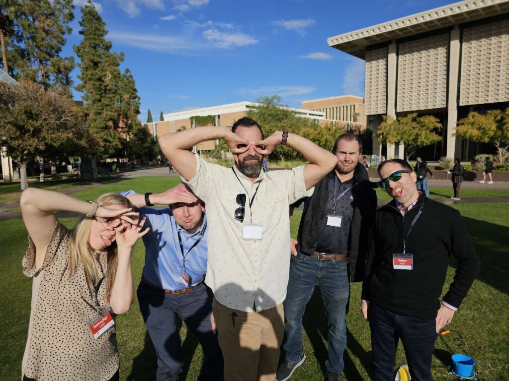 Team building participants strike playful poses while standing on a grassy area outside a building.