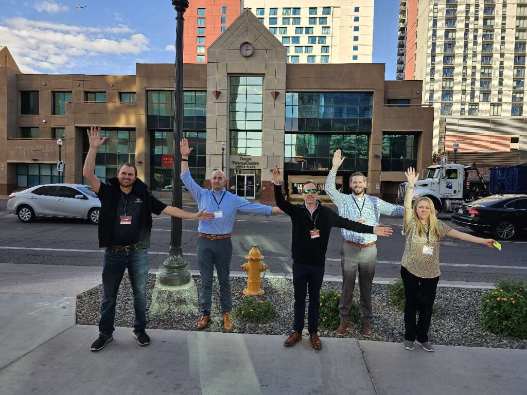 Five team members standing outside, smiling and raising their arms enthusiastically as part of a team building activity during a scavenger hunt, with an urban backdrop of buildings behind them.