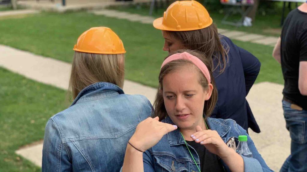 Outdoor team building exercise focused on non-verbal communication. Participants wearing hard hats use gestures to relay a message while teammates sit back-to-back, highlighting teamwork and effective information transfer.