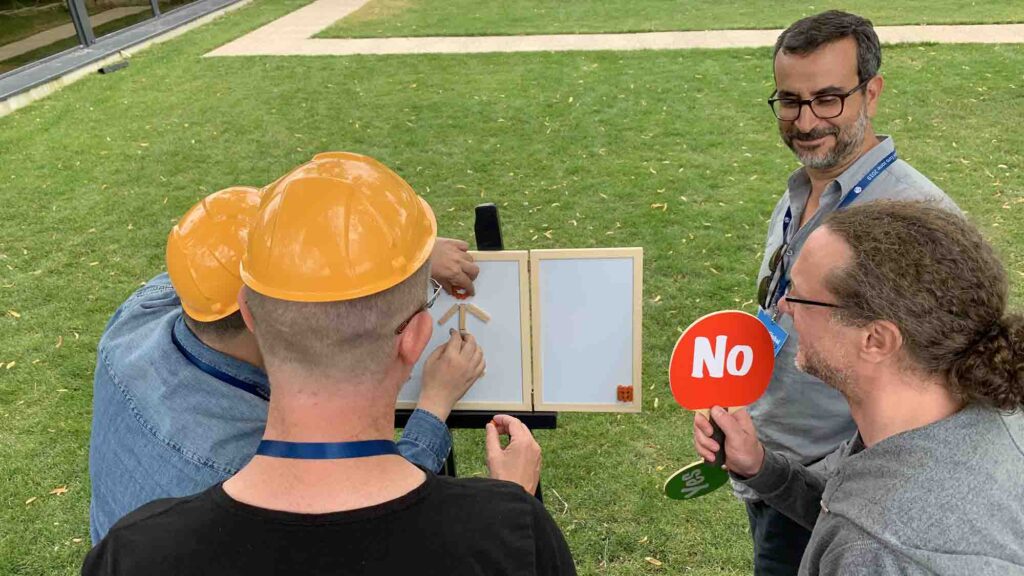 Outdoor team building activity focused on communication and feedback. Participants wearing hard hats work together to assemble a model while receiving 'yes' or 'no' responses, reinforcing teamwork and clear instruction skills.