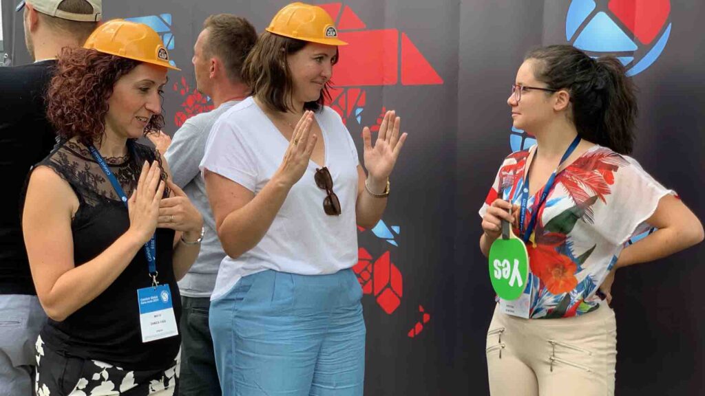 Team building activity focused on non-verbal communication. Participants wearing hard hats use gestures to convey a message while receiving feedback, emphasizing teamwork, clarity, and collaboration.