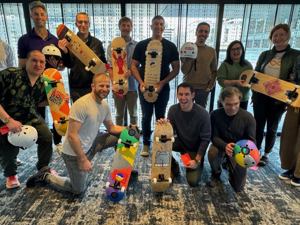 A group of people standing together proudly displaying their custom-designed skateboards and helmets. The skateboards are creatively painted with vibrant colors and designs, showcasing individuality and team spirit. Everyone is smiling, reflecting the success of their collaborative efforts in this charitable team building event.