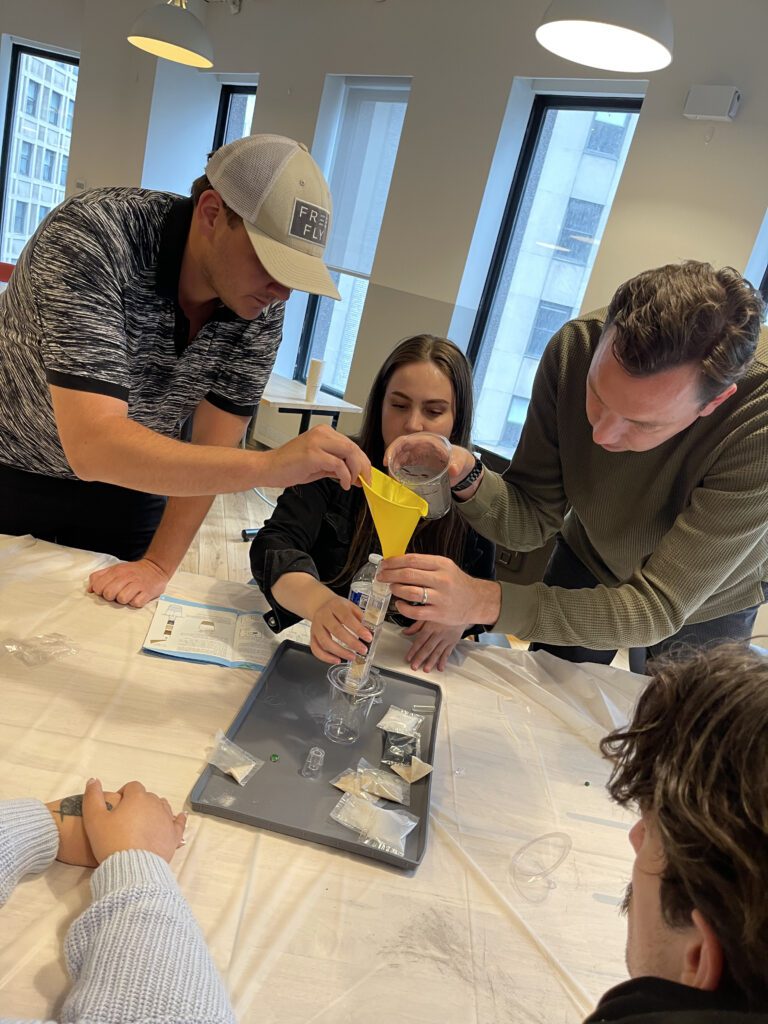 Three participants work together to pour water through a funnel into a water filtration system during a Clean Water Connection team building event. The hands-on activity teaches participants how water filtration systems work and raises awareness about the global water crisis.