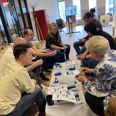 A diverse team of adults engaging in a hands-on clean water filter assembly as part of the Clean Water Connection event. The group works together in a collaborative environment, assembling water filtration kits while learning about the global water crisis. The interactive team building activity fosters community engagement.