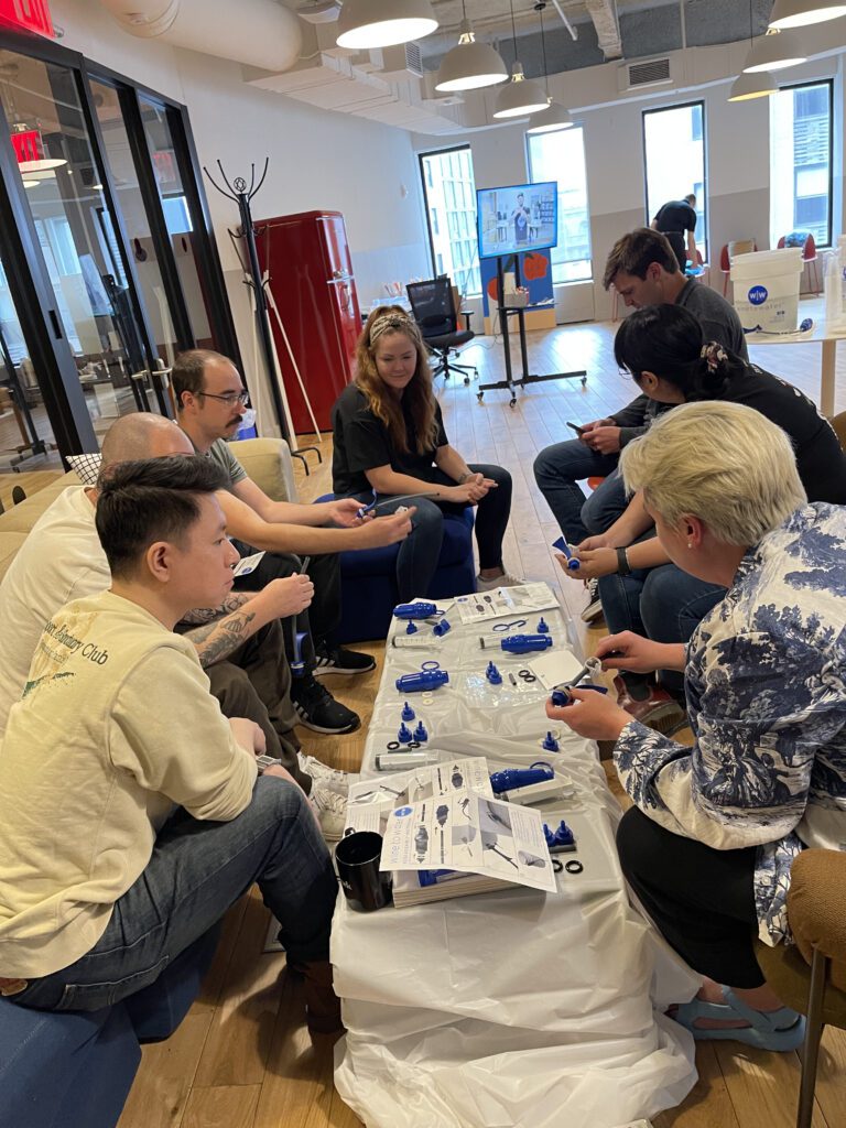 A diverse team of adults engaging in a hands-on clean water filter assembly as part of the Clean Water Connection event. The group works together in a collaborative environment, assembling water filtration kits while learning about the global water crisis. The interactive team building activity fosters community engagement.