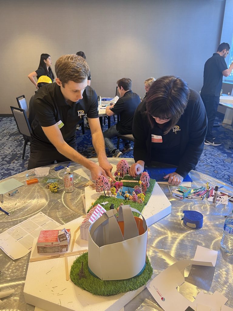 Two participants at a City Build team building event focus on constructing a detailed miniature city model using materials like trees, buildings, and flags.