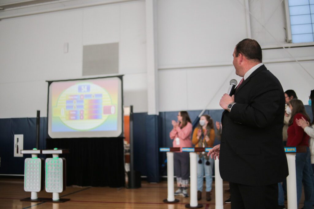 A game show host holding a microphone and looking at a large screen displaying the game board, while participants cheer and clap during the Survey Says team building activity in a gymnasium setting.