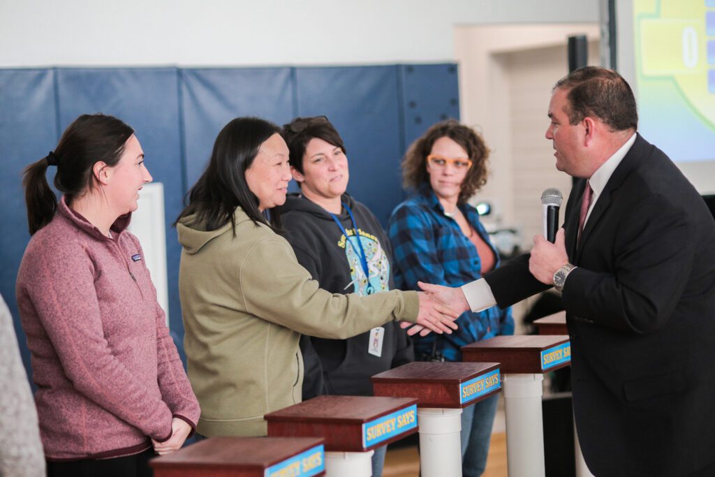 A game show host shaking hands with a participant while holding a microphone, as three other participants look on, during the Survey Says team building activity.