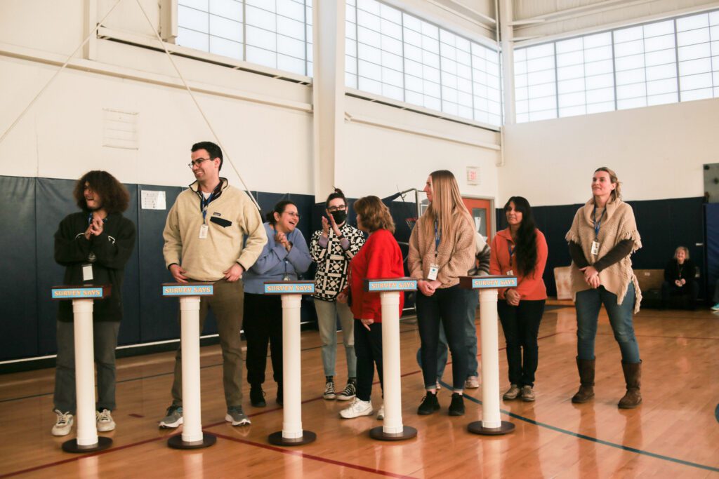 A group of participants standing behind individual podiums, ready to compete in the Survey Says team building activity, set up in a gymnasium, displaying anticipation and team spirit.