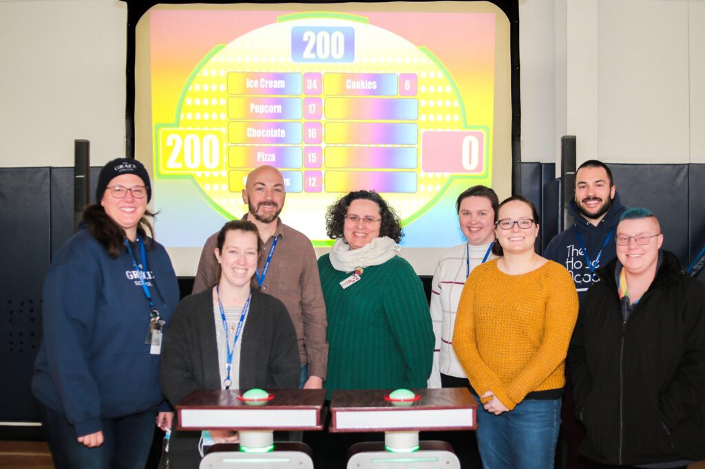 A group of eight participants smiling and standing together in front of a game show setup with a brightly colored game board in the background during the Survey Says game.