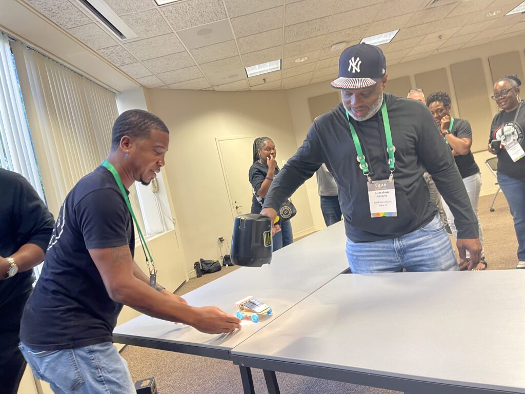 Two participants testing the performance of a small model car on a table during the STEM Speedway team building activity, with one participant using a fan to simulate wind conditions, showcasing teamwork, experimentation, and problem-solving.