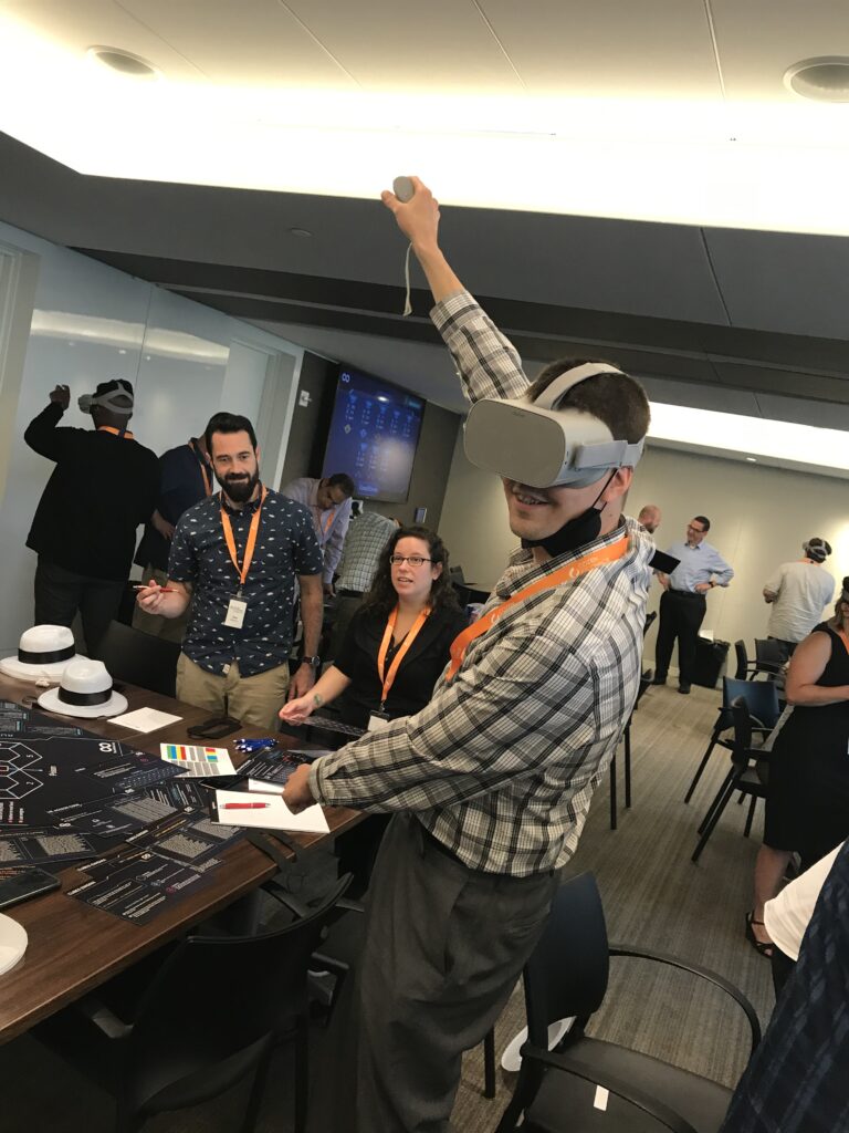 A participant wearing a VR headset raises his arm while engaging in The Infinite Loop team building activity, surrounded by teammates and facilitators in a collaborative environment.