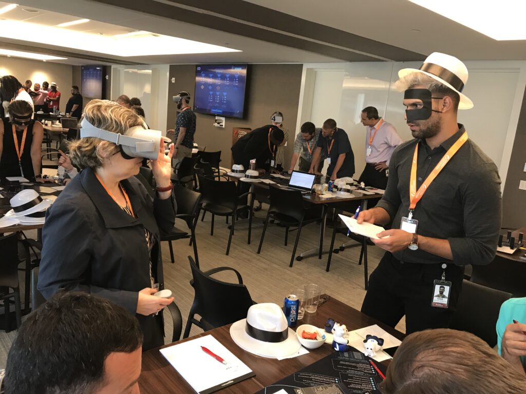 Participants at The Infinite Loop team building event engage in problem-solving activities, with one person wearing a VR headset and another taking notes while wearing a themed mask and hat.