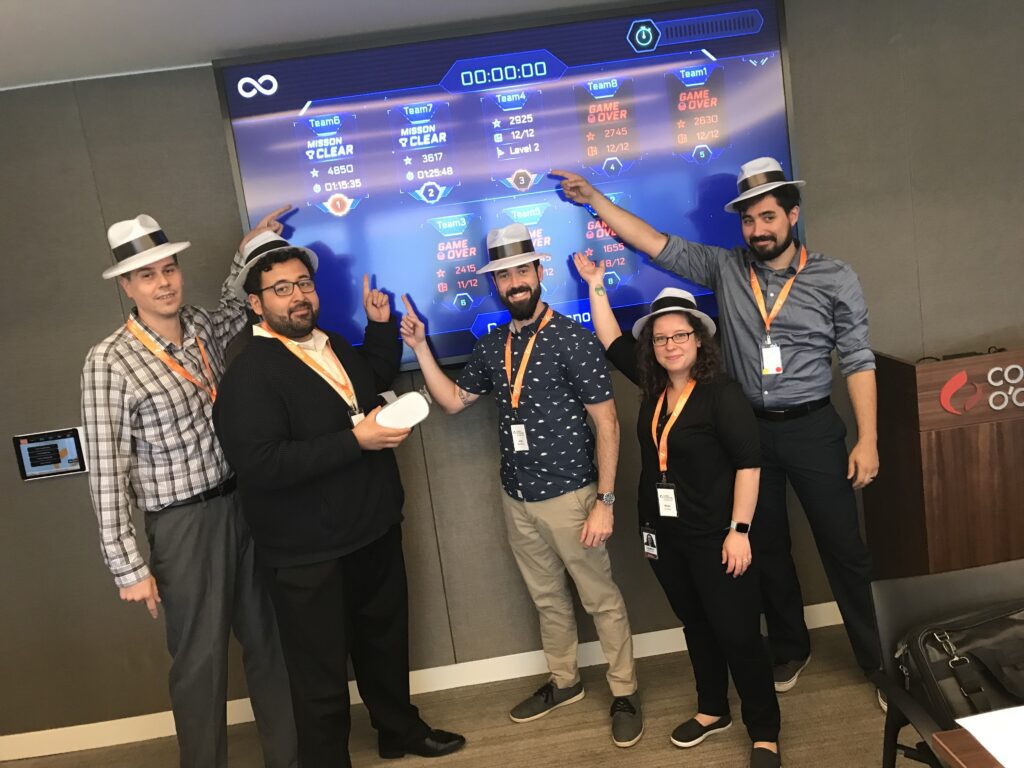 A team celebrates their progress, pointing at a virtual leaderboard during The Infinite Loop team building event, while wearing themed hats and badges.