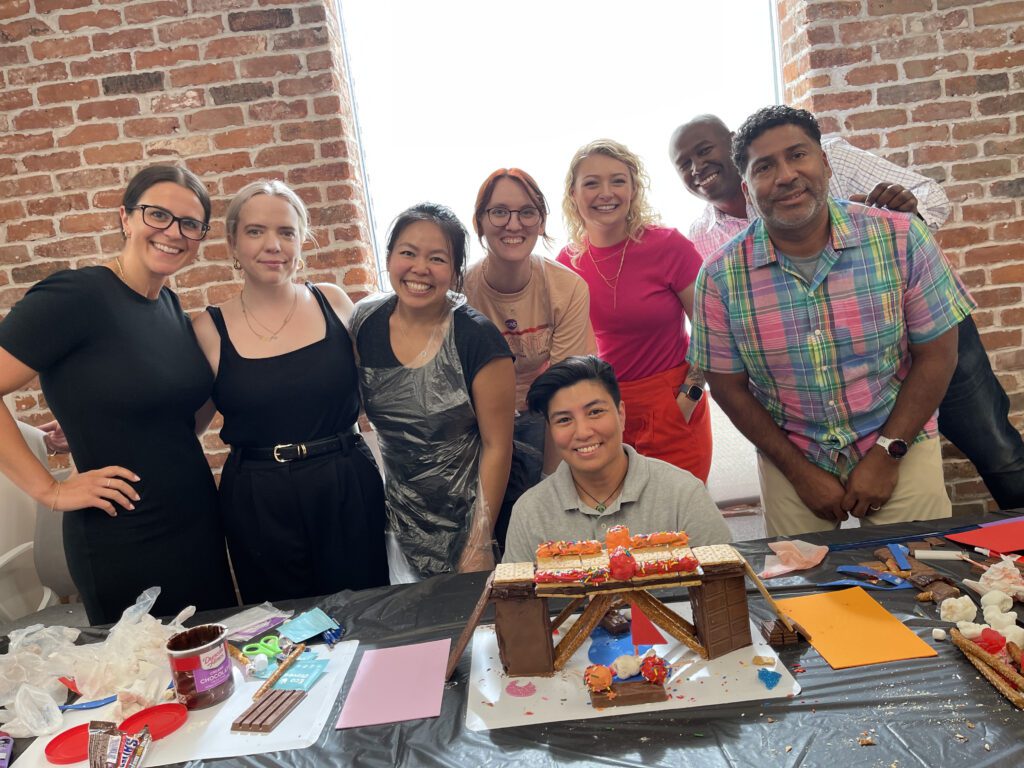 A group of smiling participants at The Chocolate Challenge team building event stands proudly behind their edible chocolate bridge creation. The table in front of them is covered with supplies like frosting, sprinkles, and candy, used to design their structure.