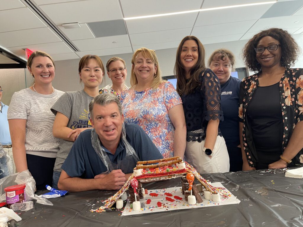 A group of smiling participants stands behind their completed chocolate bridge at The Chocolate Challenge team building event.