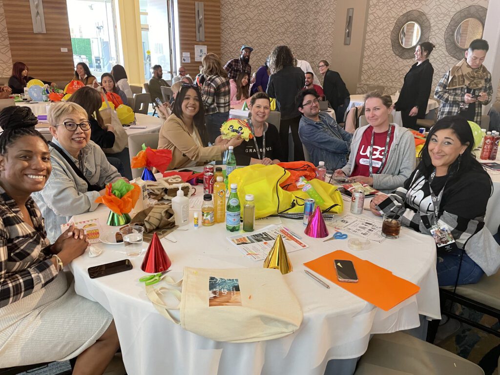 A group of people seated around a table, smiling and interacting, with colorful party hats, birthday-themed decorations, and supplies spread out, engaging in a team building activity for a charitable cause.