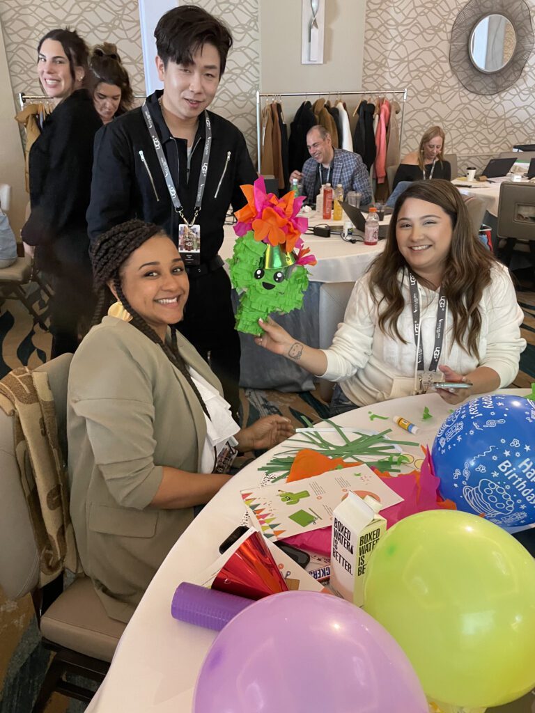 Two women smile while holding up a green cactus-themed piñata decorated with colorful flowers, sitting at a table filled with birthday craft supplies and party decorations. Balloons add to the celebratory mood of this charitable team building program.