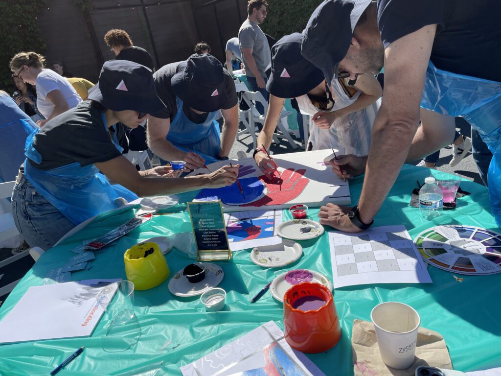 A group of participants in matching hats and aprons collaborate on painting a vibrant section of a mural during The Big Picture team building event. They work together, adding colors to the canvas under a sunny outdoor setting, with painting supplies and color wheels scattered on the table.