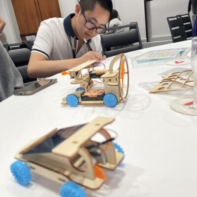 A participant focused on assembling a miniature car from a STEM kit during the STEM Speedway team building activity, with completed wooden model cars on the table, highlighting precision, creativity, and hands-on learning.