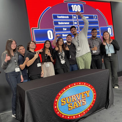 A group of participants smiling and giving thumbs up in front of a large game board during the Survey Says team-building activity, celebrating their participation in a fun, competitive, and interactive game.