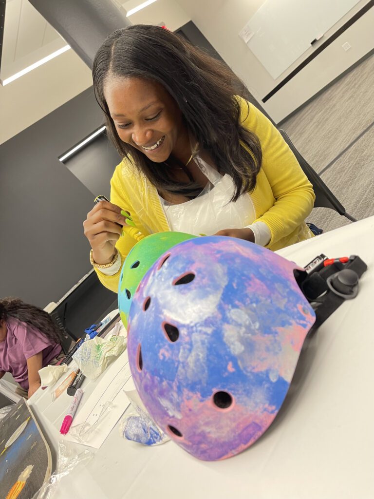 A woman with long black hair, wearing a yellow sweater, is happily painting and decorating a helmet during a team building event.