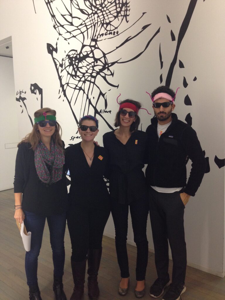 Team members in creative costumes during a museum scavenger hunt. Wearing fun glasses and engaging with an art exhibit, they work together to solve clues in this interactive team building activity.