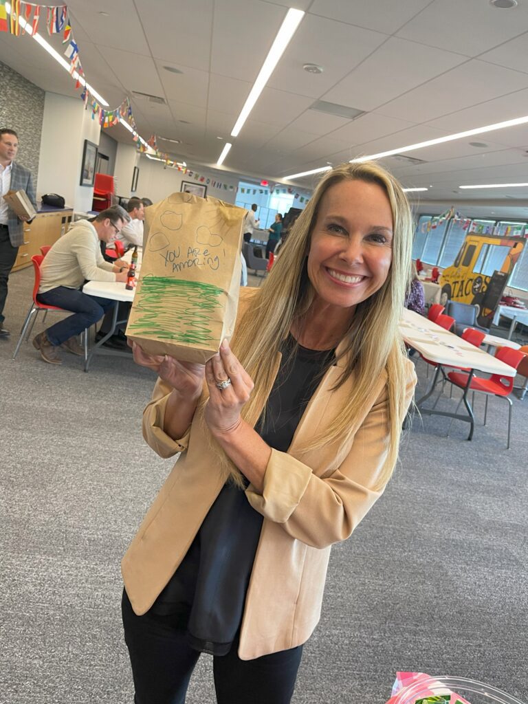 Smiling woman holding a decorated brown paper bag with the message 'You Are Amazing!' during The Great Snack Pack activity at a corporate charitable team building event.